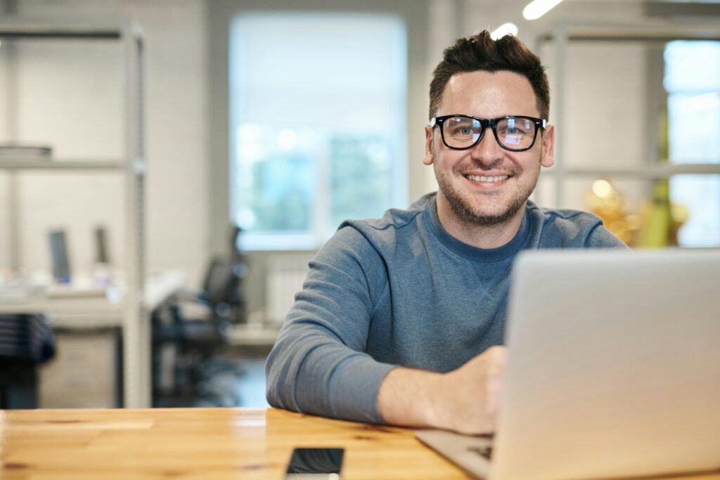 Happy man wearing glasses working remotely on laptop in modern office environment.